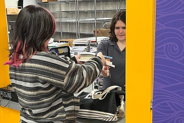 A woman with pink streaks in her dark hair who is wearing a multicolored striped sweater and cargo pants faces a counter in Dowdy Student Stores where she is picking up several books from a female employee with dark shoulder length hair wearing a gray shirt. 