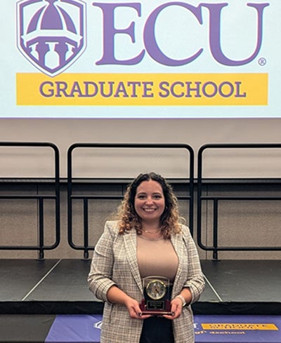 A woman in dark slacks, tan shirt and a gray plaid blazer holds a first-place trophy and stands in front of a table with a tablecloth advertising the Three-Minute Thesis competition.