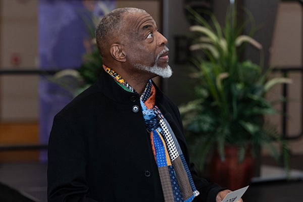 A nearly bald man with a white mustache and goatee, wearing a colorful scarf and black suit jacket, holds a open card with the ECU logo on the front as he looks up at the space of the Main Campus Student Center ballroom.