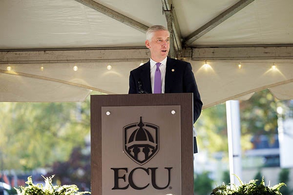 ECU’s chancellor stands and speaks into a microphone at a tall, wooden podium with the ECU logo/crest displayed on the front.