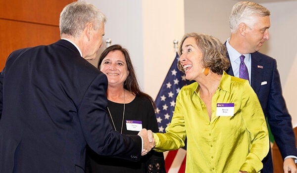 A woman in a bright green shirt with light brown hair shakes the hand of a man in a suit while another woman dressed in black with dark hair looks on.